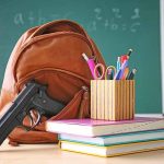 Gun and backpack on desk with school supplies.