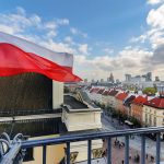 Polish flag over cityscape with buildings and cloudy sky.