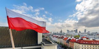 Polish flag over cityscape with buildings and cloudy sky.