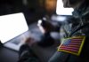 Soldier using laptop with US flag patch visible.