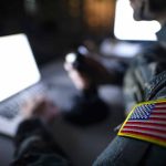 Soldier using laptop with US flag patch visible.