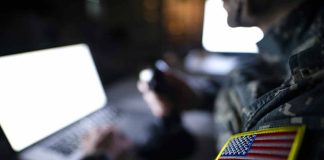 Soldier using laptop with US flag patch visible.