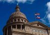 Building dome with US and Texas flags, blue sky.