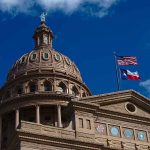 Building dome with US and Texas flags, blue sky.