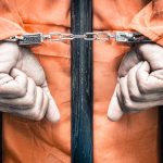 Handcuffed hands in orange jumpsuit behind bars.