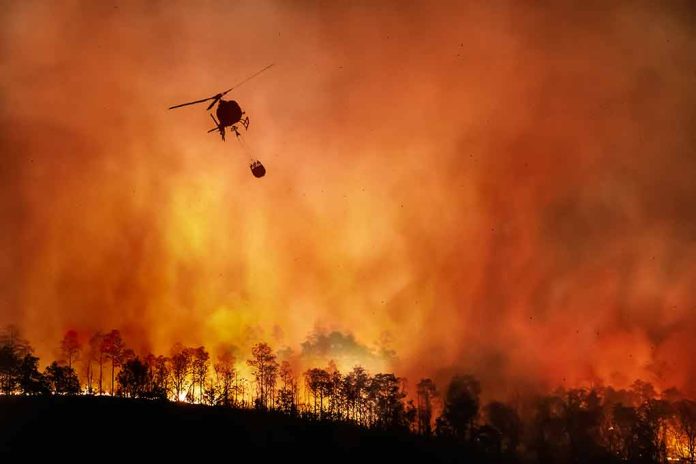 Helicopter fighting massive forest fire with water bucket.