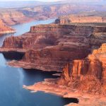 Red rock canyons beside blue water body.