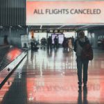 Airport sign: "All Flights Canceled", passengers walking.