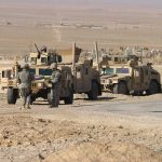 Soldiers standing near armored vehicles in desert terrain.