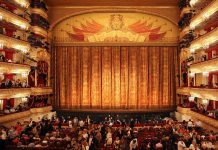 Ornate theater, grand curtain, audience seated below balconies.