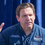 Man speaking, wearing a blue "Desantis for President" shirt.