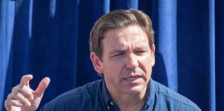 Man speaking, wearing a blue "Desantis for President" shirt.