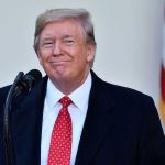 Trump Charms PM – Lands MAJOR Deal! Man in suit and tie speaking at podium.