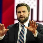 Man in suit gesturing with hands on stage.