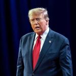 Trump SUFFERS GAFFE – Serious Blank Moment! Man in suit with red tie speaking on stage.