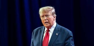 Trump SUFFERS GAFFE – Serious Blank Moment! Man in suit with red tie speaking on stage.