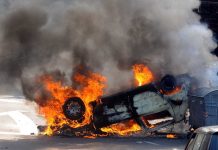 An overturned car engulfed in flames and smoke on a city street