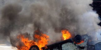 An overturned car engulfed in flames and smoke on a city street