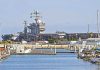 Trump DEPLOYS USS Ford – See What’s Brewing A large naval aircraft carrier docked in a harbor with smaller boats in the foreground
