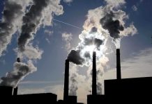 Silhouetted smokestacks emitting smoke against a cloudy sky with sunlight