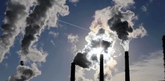 Silhouetted smokestacks emitting smoke against a cloudy sky with sunlight