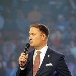 Socialist Drift Rumors Rattle Richmond man in a suit speaking into a microphone on stage