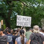 Rushed Settlements? DOJ Under Fire Crowd holds No More Mandates sign at protest.