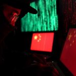 25,000 Chinese Spies Hidden Among Us? Hooded person using a laptop with Chinese flags on screens
