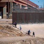 Workers Purged, Foreign Replacements Welcomed People walking along a dry canal beside a high fence with barbed wire