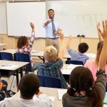 Teacher at front of classroom as students raise hands