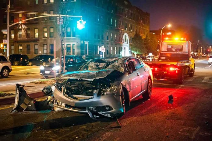 Wrecked silver car on city street at night with tow truck nearby